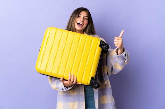 Young caucasian woman isolated on purple in vacation with travel suitcase and with thumb up