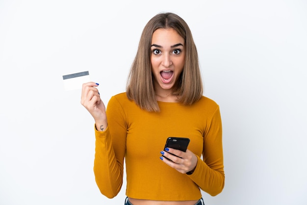 Young caucasian woman isolated on white background buying with the mobile and holding a credit card with surprised expression