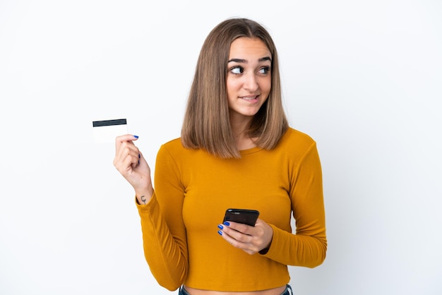 Young caucasian woman isolated on white background buying with the mobile with a credit card while thinking