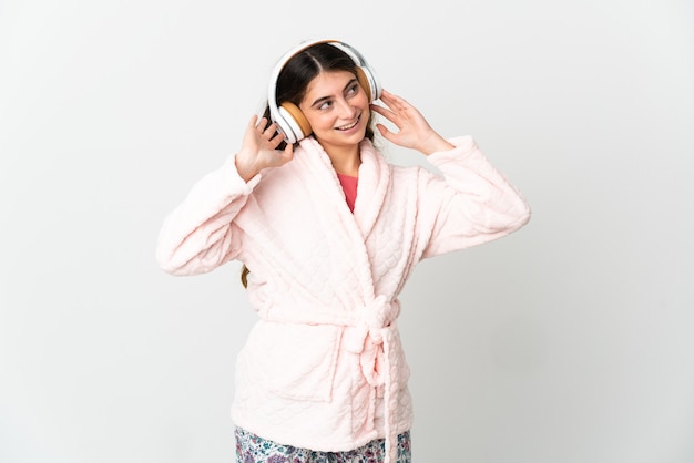 Young caucasian woman isolated on white background in pajamas and holding a pillow and listening music