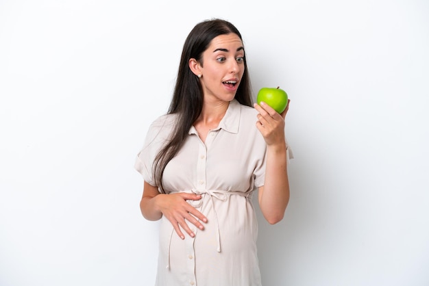 Young caucasian woman isolated on white background pregnant and holding an apple