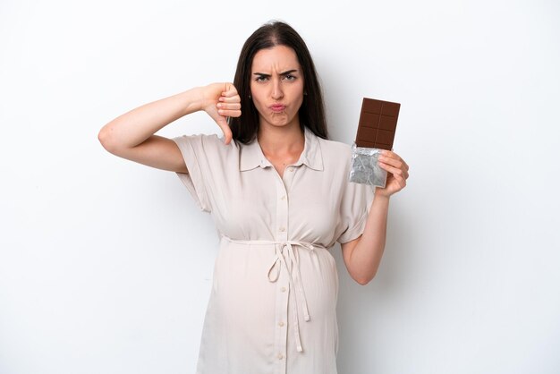 Young caucasian woman isolated on white background pregnant and holding chocolate doing bad signal