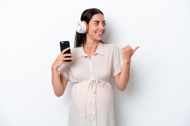 Young caucasian woman isolated on white background pregnant and listening music while pointing side