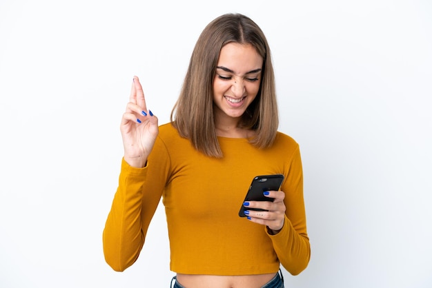 Young caucasian woman isolated on white background using mobile phone with fingers crossing