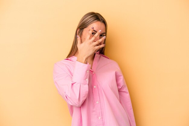 Young caucasian woman isolated on yellow background blink at the camera through fingers, embarrassed covering face.