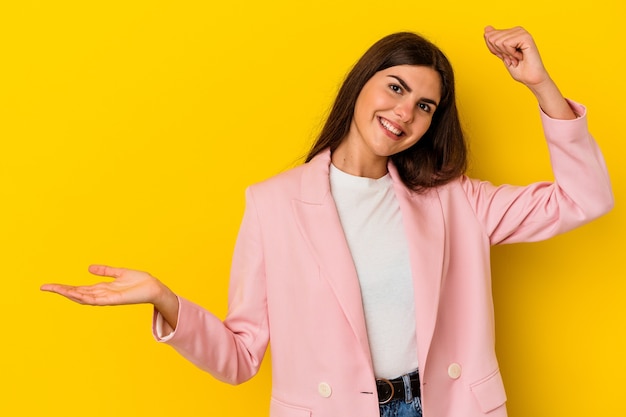 Young caucasian woman isolated on yellow background holds copy space on a palm, keep hand over cheek. Amazed and delighted.