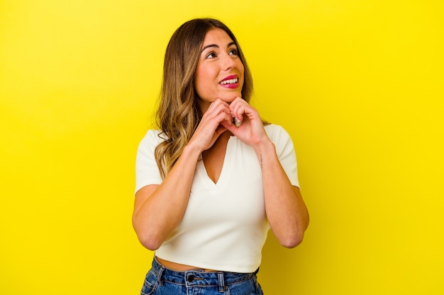 Young caucasian woman isolated on yellow background keeps hands under chin, is looking happily aside.