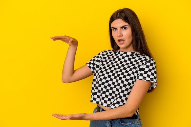 Young caucasian woman isolated on yellow background shocked and amazed holding a copy space between hands.