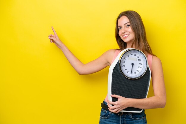 Young caucasian woman isolated on yellow background with weighing machine and pointing side