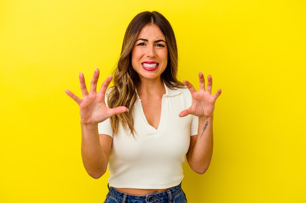 Young caucasian woman isolated on yellow wall showing claws imitating a cat, aggressive gesture.