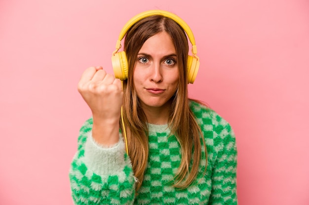 Young caucasian woman listening to music isolated on pink background showing fist to camera aggressive facial expression