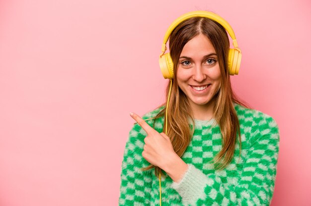 Young caucasian woman listening to music isolated on pink background smiling and pointing aside showing something at blank space