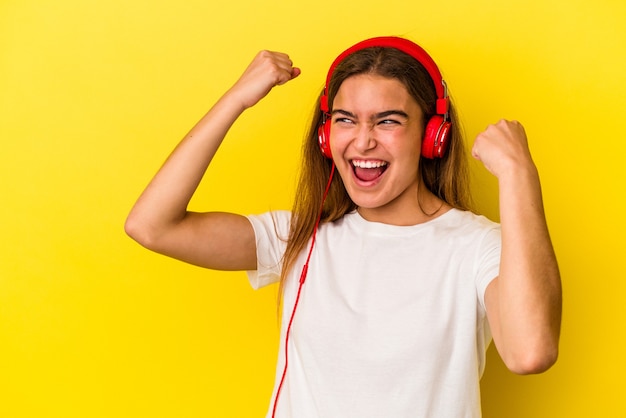 Young caucasian woman listening to music isolated on yellow background raising fist after a victory, winner concept.