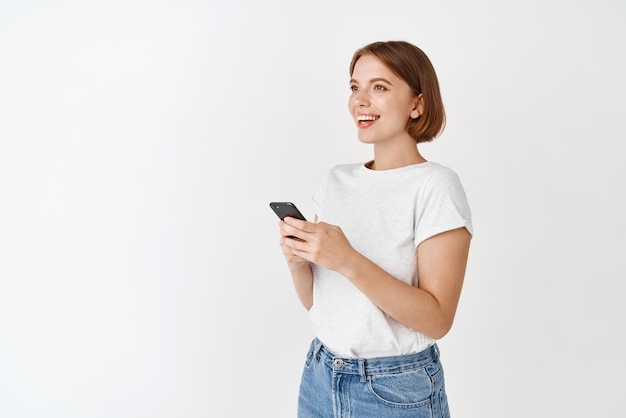 Young caucasian woman looking aside at logo holding cell phone in hands Happy girl chatting in app on smartphone standing in tshirt with jeans white background