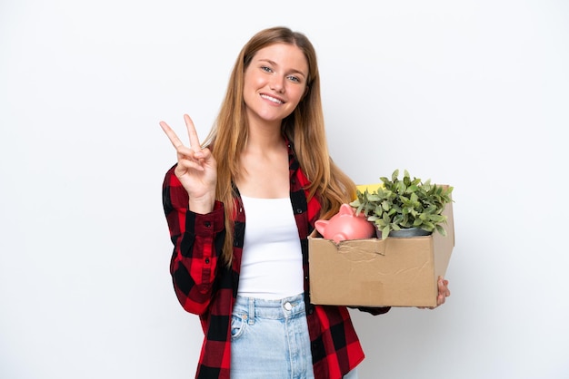 Young caucasian woman making a move while picking up a box full of things isolated on white background smiling and showing victory sign