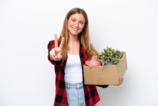 Young caucasian woman making a move while picking up a box full of things isolated on white background smiling and showing victory sign