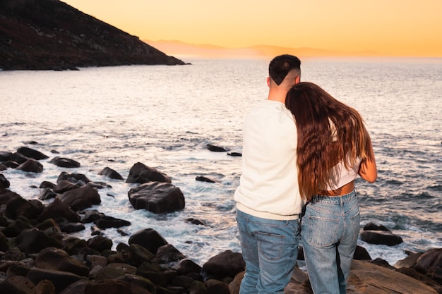 Young caucasian woman and man embracing each other at a rocky beach while sunset