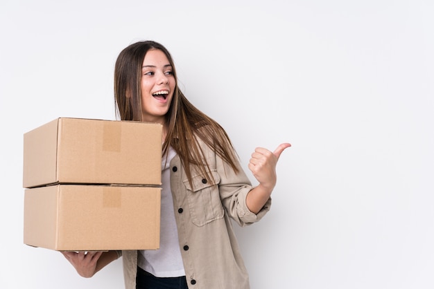 Young caucasian woman moving to a new home points with thumb finger away, laughing and carefree