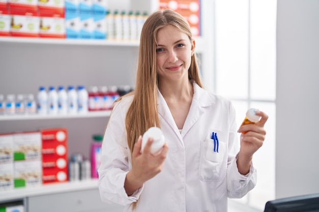 Young caucasian woman pharmacist smiling confident holding pills bottles at pharmacy