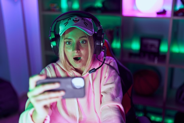 Young caucasian woman playing video games with smartphone pointing thumb up to the side smiling happy with open mouth