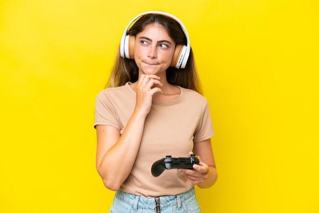 Young caucasian woman playing with a video game controller isolated on yellow background having doubts and thinking