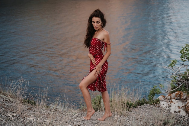 A young Caucasian woman poses in a red dress at the edge of a cliff near the beach and the sea