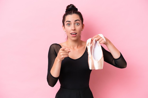 Young caucasian woman practicing ballet isolated on pink background surprised and pointing front