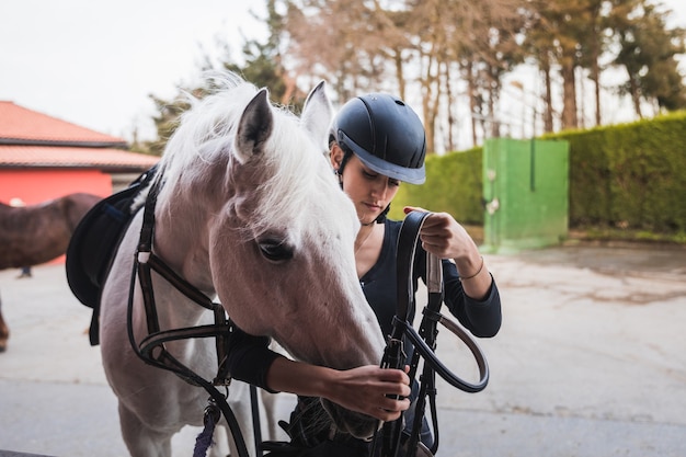 Young caucasian woman preparing a white horse for a ride