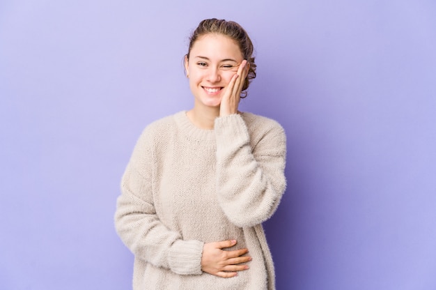 Young caucasian woman on purple wall laughs happily and has fun keeping hands on stomach.