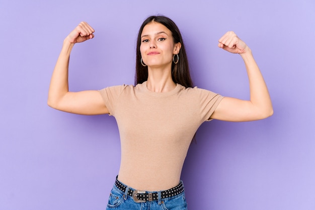 Young caucasian woman on purple wall showing strength gesture with arms, symbol of feminine power