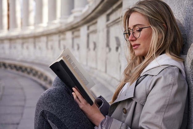 Young caucasian woman reading book in park with antique building