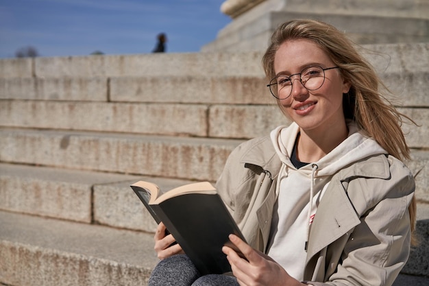 Young caucasian woman reading book in park with antique building