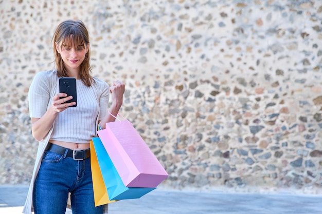 Young caucasian woman shopping with bags in one hand looking at her smart phone in the street