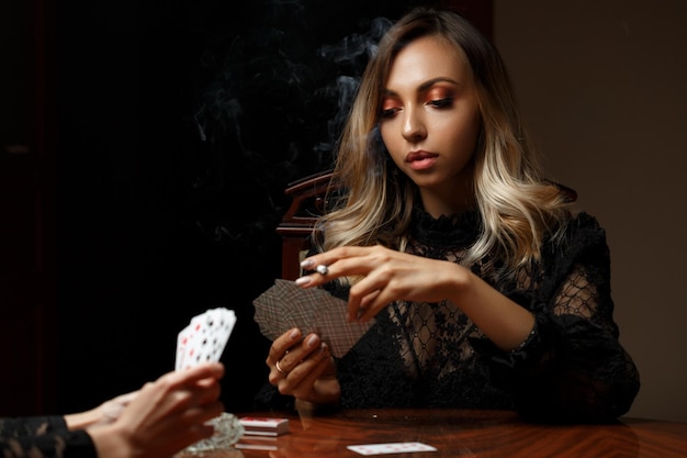 Young caucasian woman sits at a table plays cards for undressing and smokes