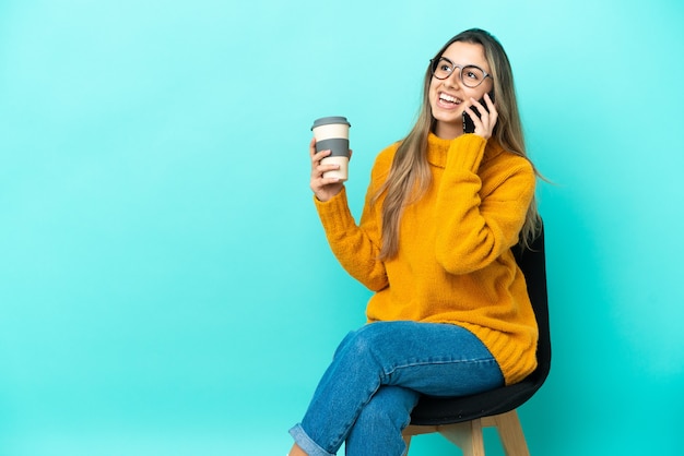 Young caucasian woman sitting on a chair isolated on blue background holding coffee to take away and a mobile