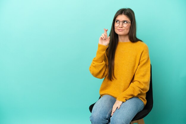 Young caucasian woman sitting on a chair isolated on blue background with fingers crossing and wishing the best