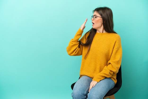 Young caucasian woman sitting on a chair isolated on blue background yawning and covering wide open mouth with hand