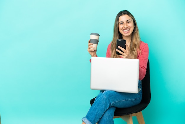 Young caucasian woman sitting on a chair with her pc isolated on blue background holding coffee to take away and a mobile