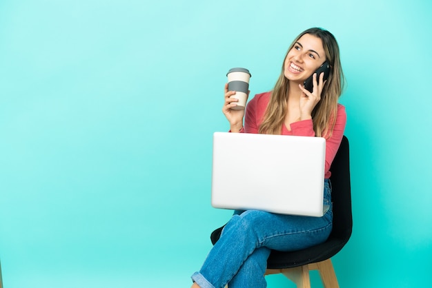 Young caucasian woman sitting on a chair with her pc isolated on blue wall holding coffee to take away and a mobile