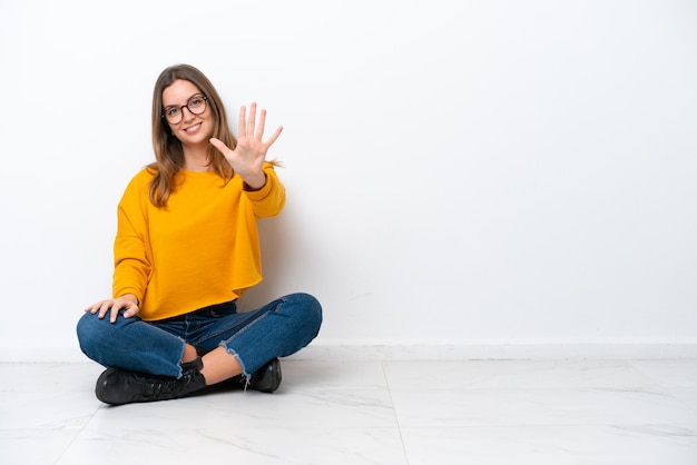 Young caucasian woman sitting on the floor isolated on white background counting five with fingers