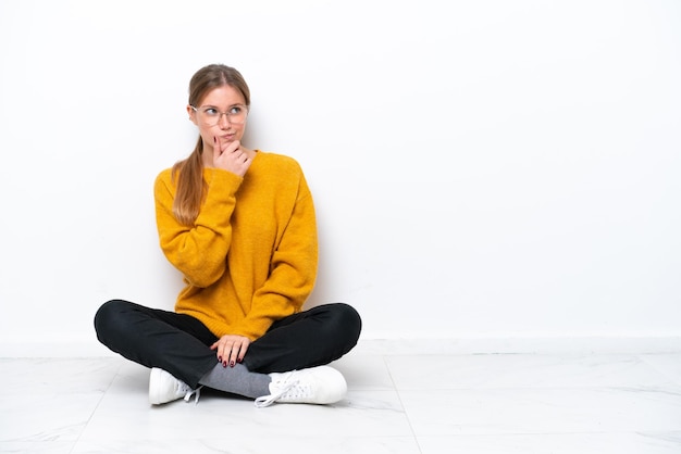 Young caucasian woman sitting on the floor isolated on white background having doubts and with confuse face expression