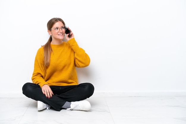 Young caucasian woman sitting on the floor isolated on white background keeping a conversation with the mobile phone