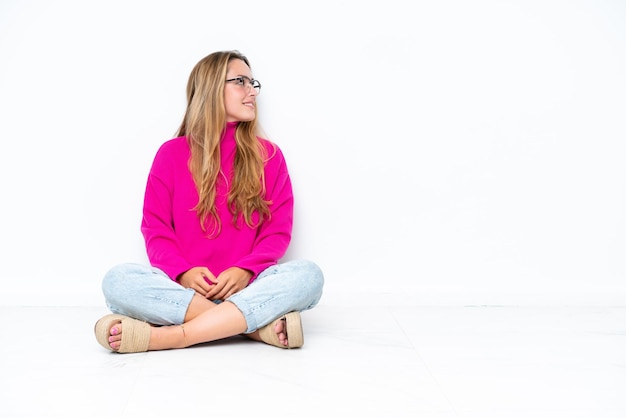 Young caucasian woman sitting on the floor isolated on white background looking side