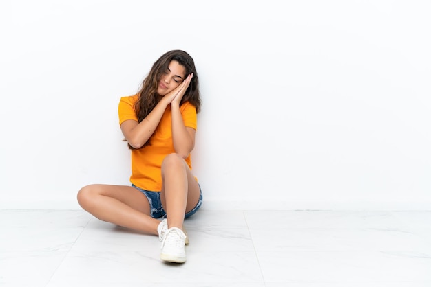 Young caucasian woman sitting on the floor isolated on white background making sleep gesture in dorable expression