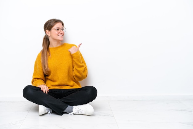 Young caucasian woman sitting on the floor isolated on white background pointing to the side to present a product