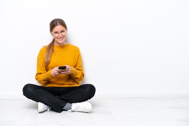 Young caucasian woman sitting on the floor isolated on white background sending a message with the mobile