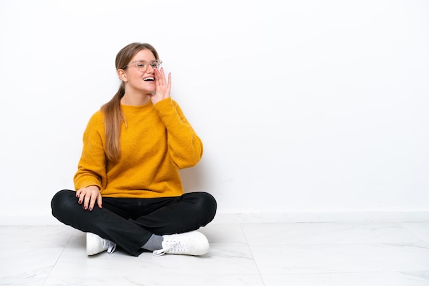 Young caucasian woman sitting on the floor isolated on white background shouting with mouth wide open to the lateral