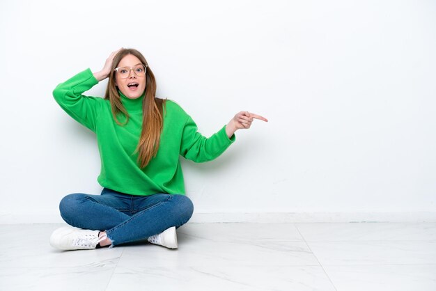 Young caucasian woman sitting on the floor isolated on white background surprised and pointing finger to the side