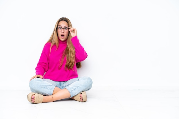 Young caucasian woman sitting on the floor isolated on white background with glasses and surprised