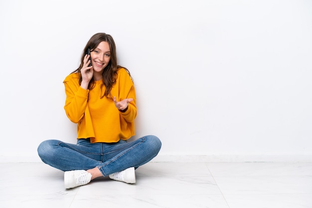 Young caucasian woman sitting on the floor isolated on white wall keeping a conversation with the mobile phone with someone